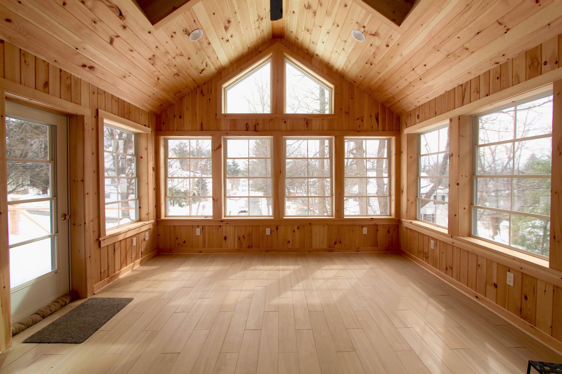 Bright wood-paneled cabin interior with large windows and vaulted ceiling, empty and sunlit.