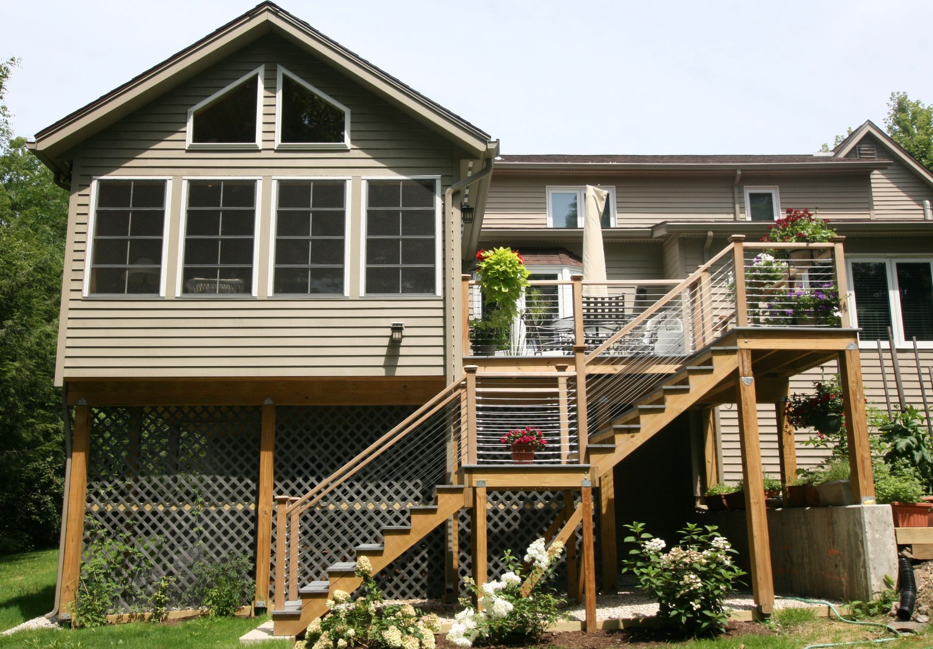 Raised house with beige siding, large windows, and a wooden staircase leading to a deck in a leafy yard