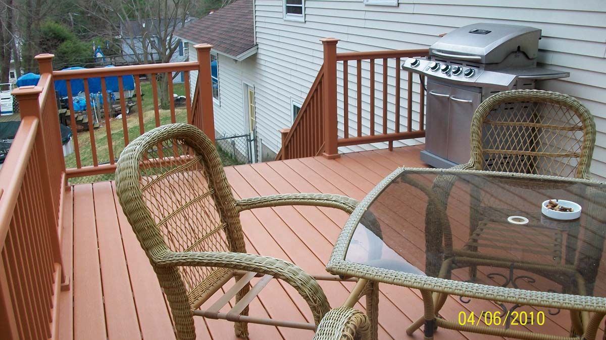 Deck with wicker patio chairs, glass table, and a grill overlooking a yard and houses.