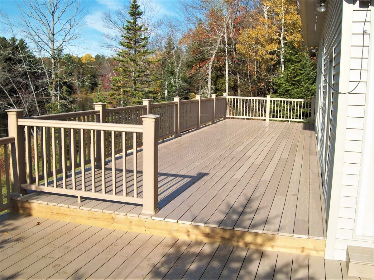 Wooden deck with railings beside a white house, overlooking trees under a clear blue sky