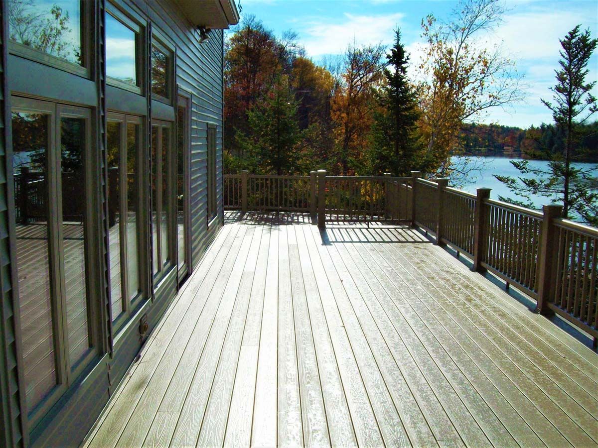 Sunlit wooden deck with railing overlooking a lake and autumn trees beside a glass house