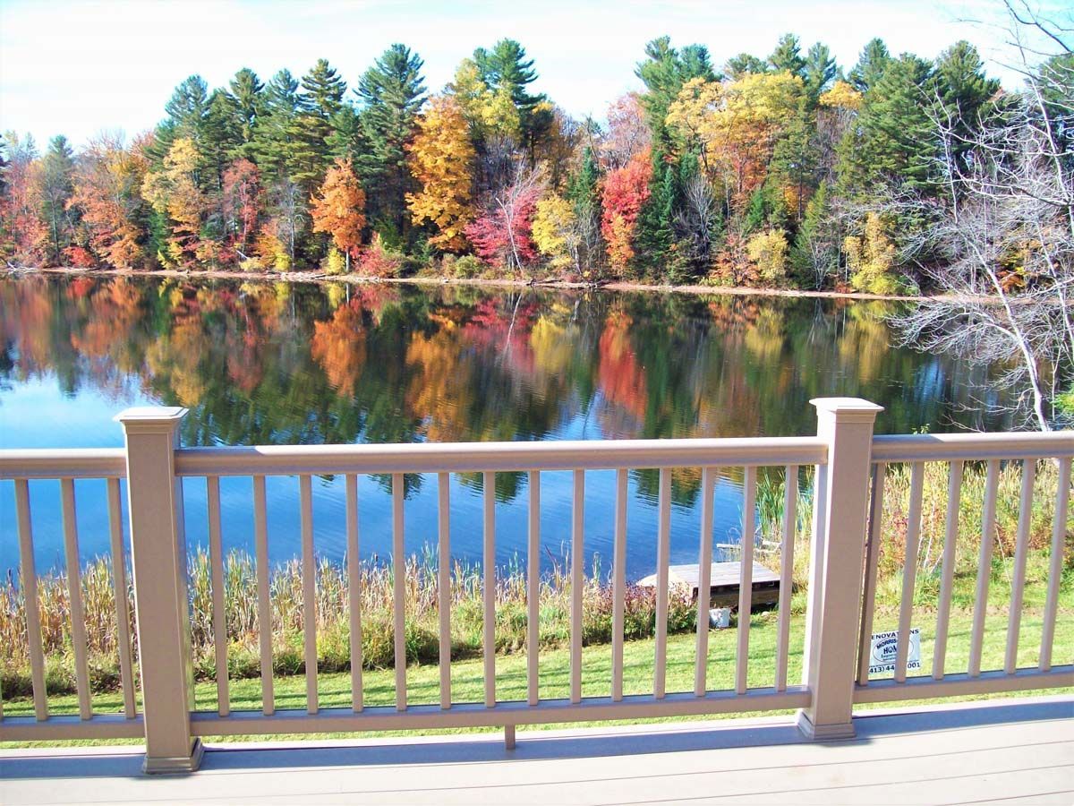 Autumn trees reflected in a calm lake, viewed from a deck with a beige railing