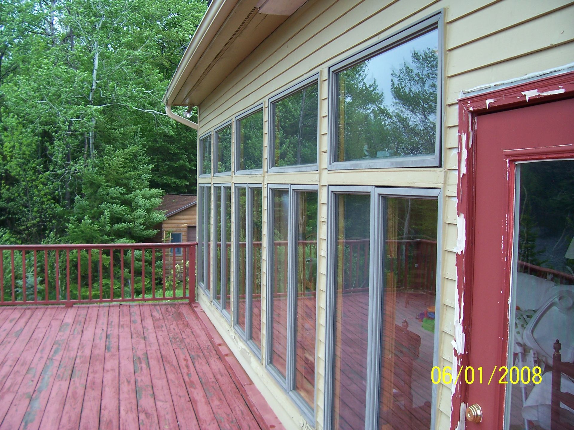 Wooden deck beside a house with large windows, surrounded by trees and a red railing.