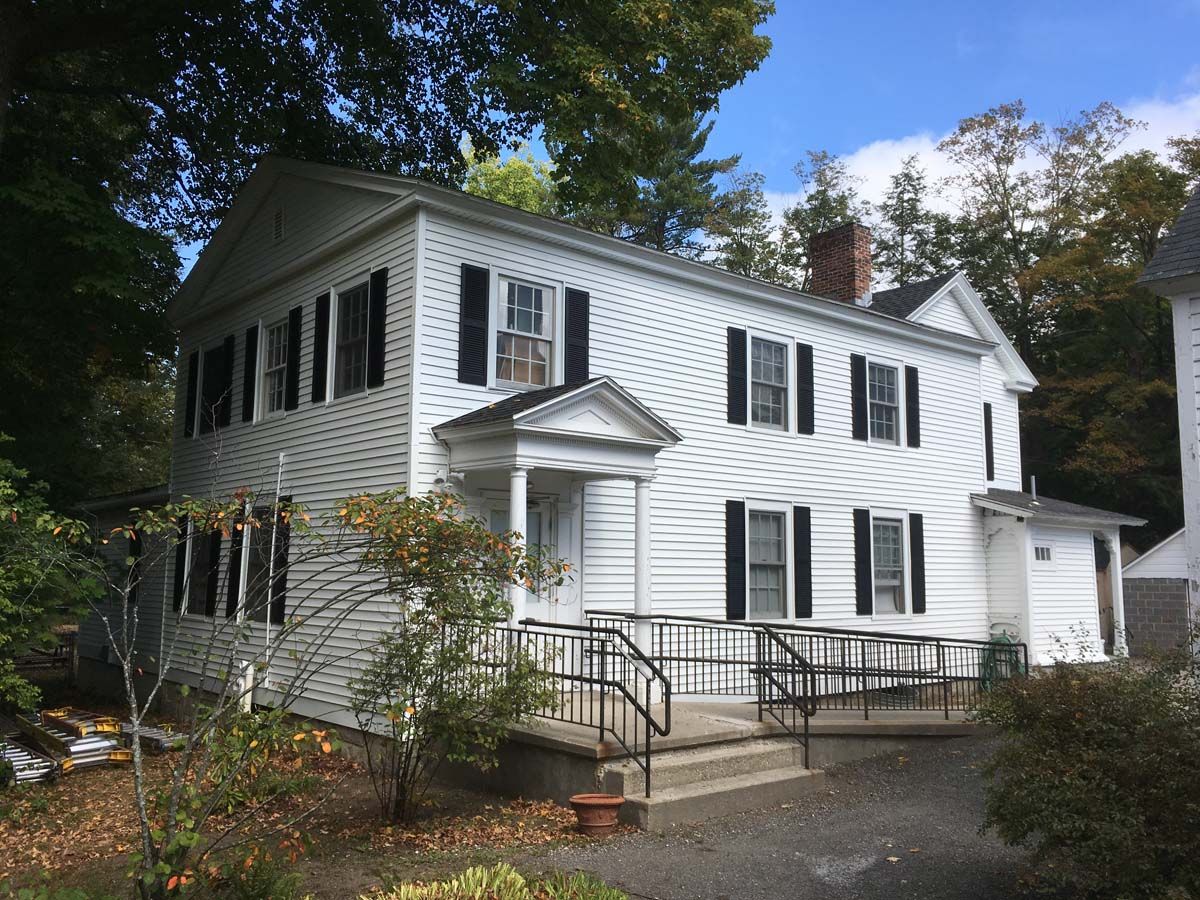 White two-story house with black shutters, front porch, and wheelchair ramp surrounded by trees