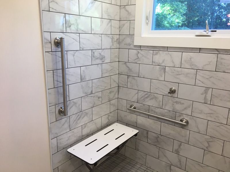 Accessible shower with tiled walls, grab bars, fold-down seat, and a small window.