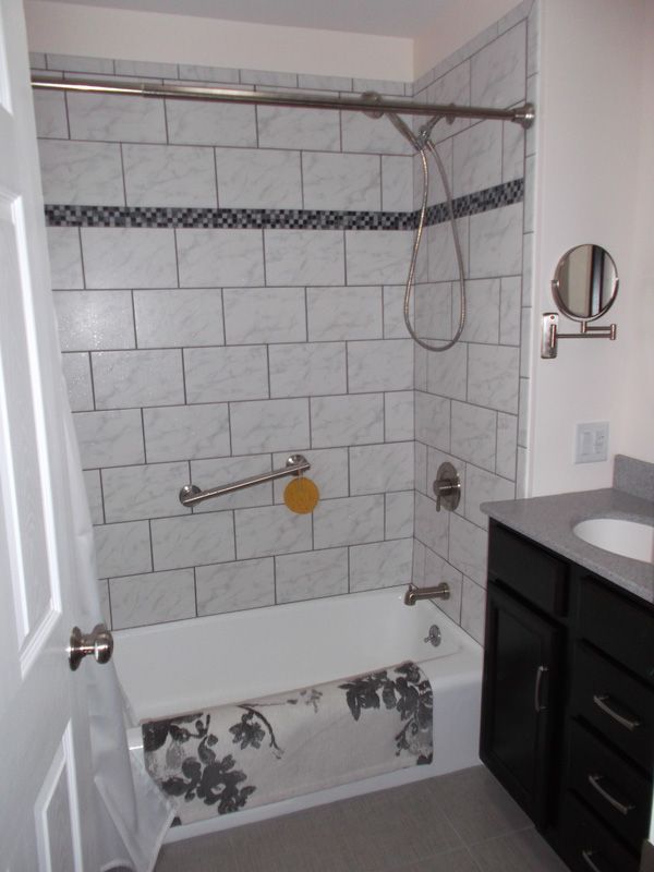 Small bathroom with a white tub, tiled walls, dark vanity, and wall-mounted shower fixtures.