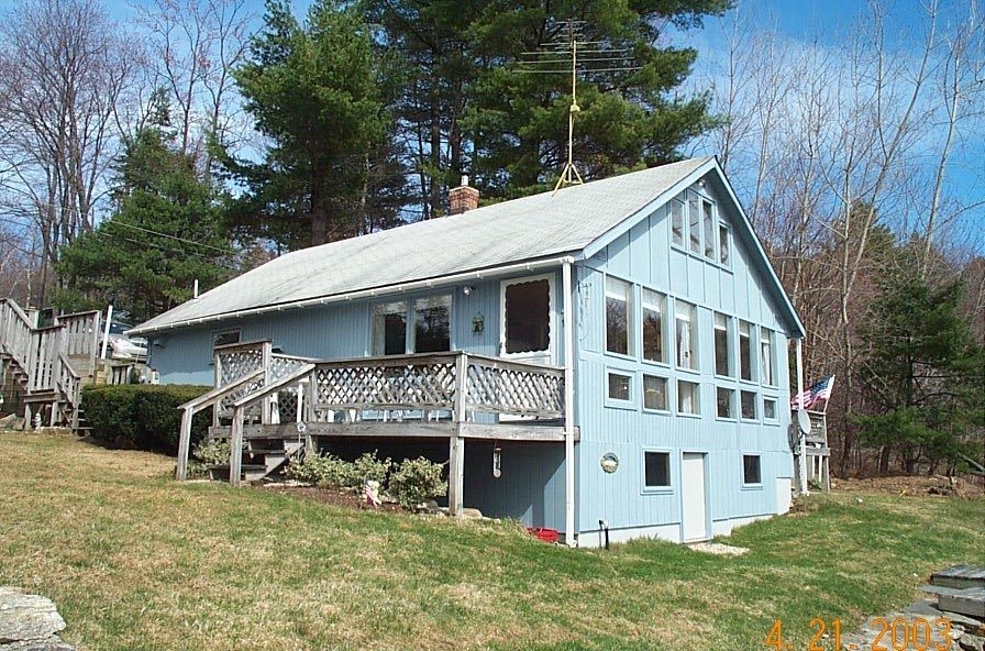 Light blue house with a porch and sloped roof on a grassy hill surrounded by trees