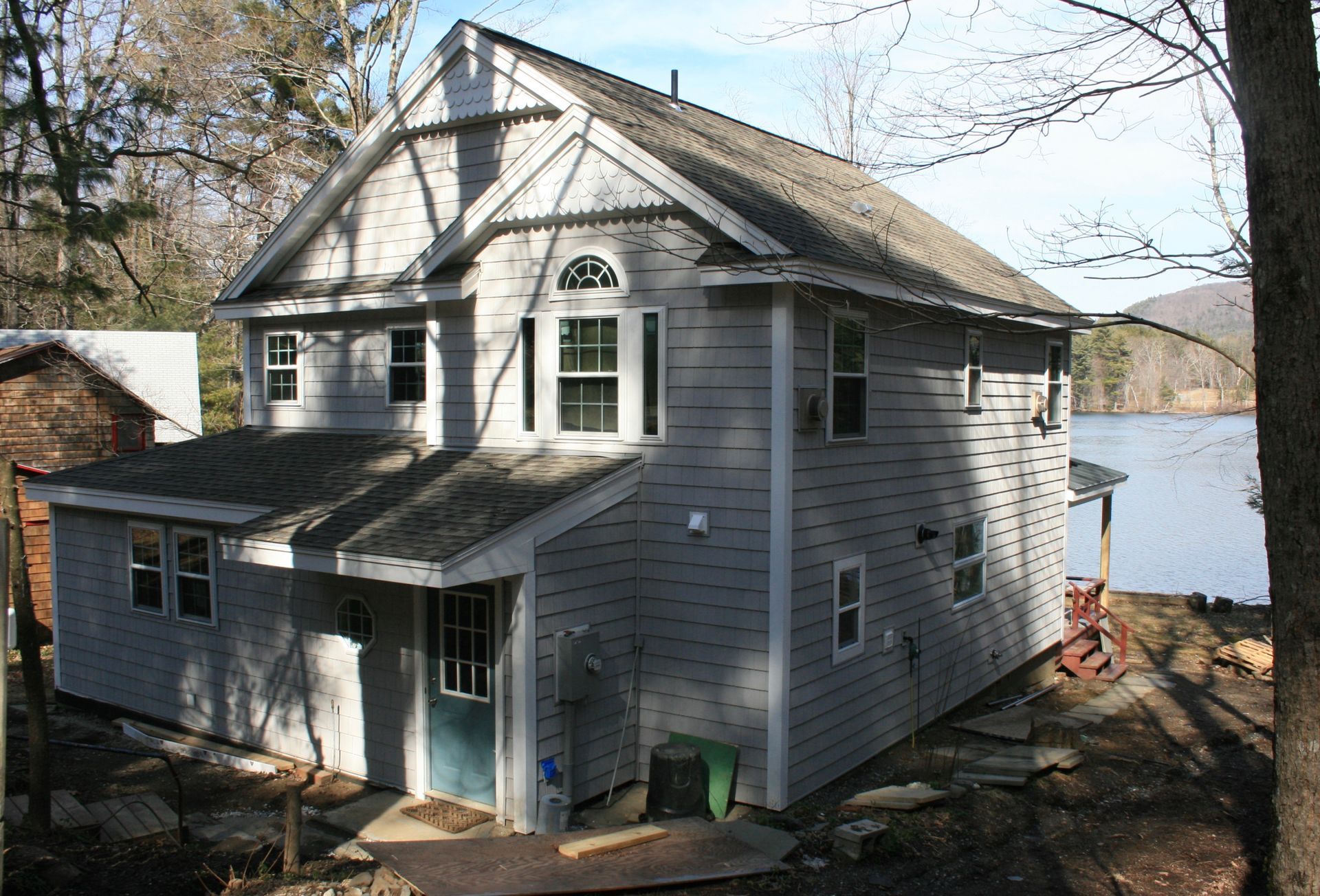 Gray lakeside house with dormers and porch, surrounded by trees under a bright sky