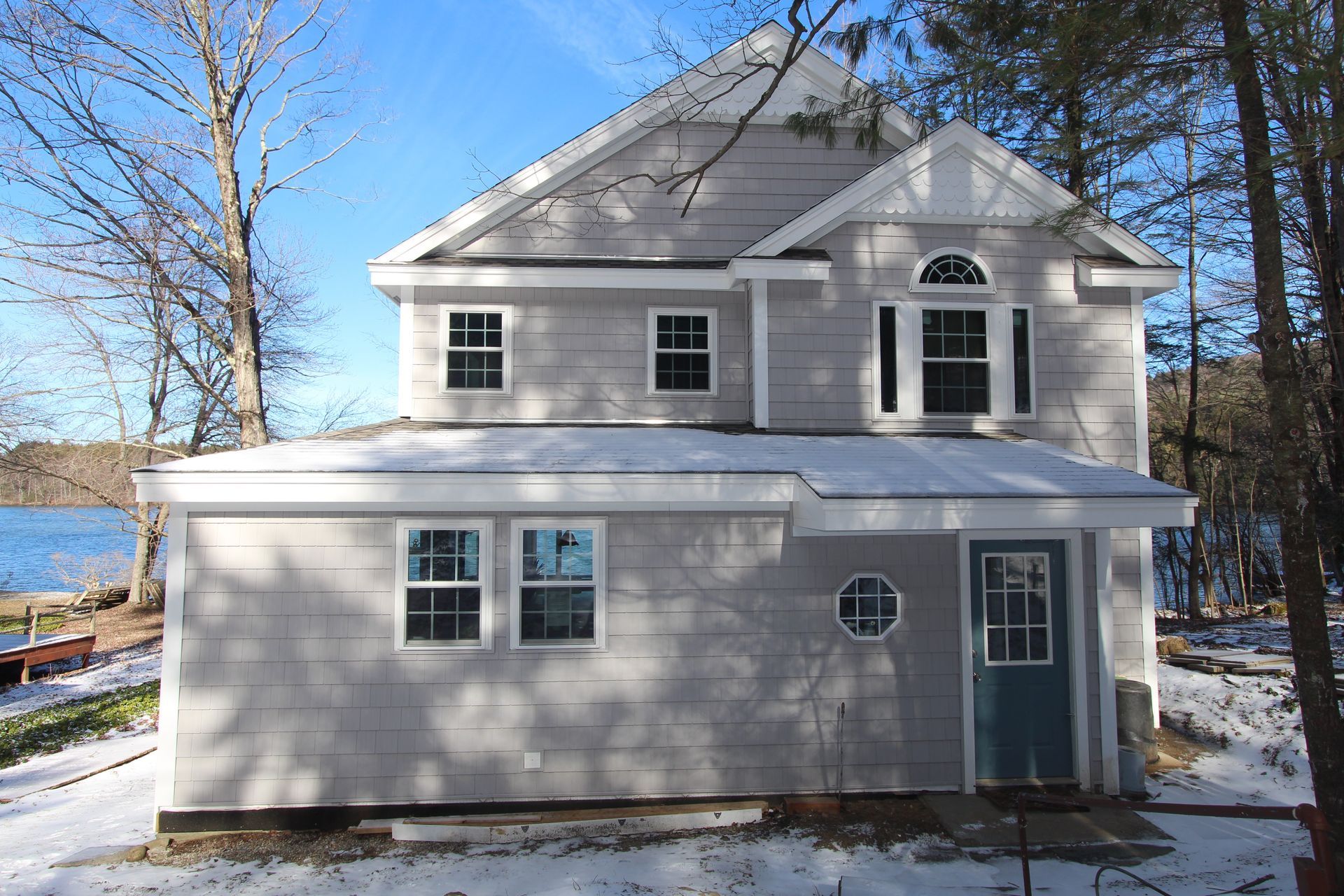 Gray two-story house with white trim and snow on the ground beside a lake.