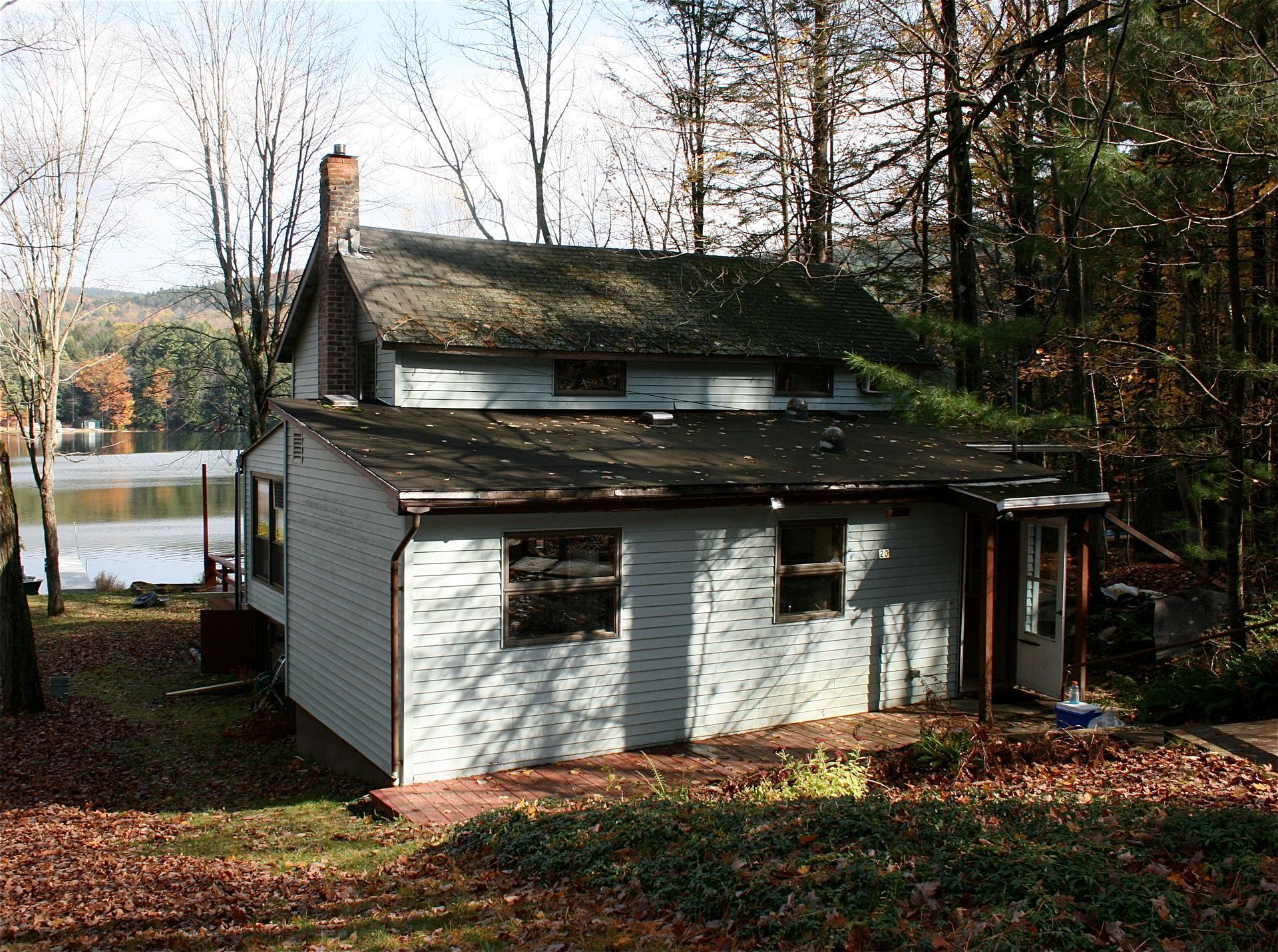 Small white house with a mossy roof in a wooded yard near a lake