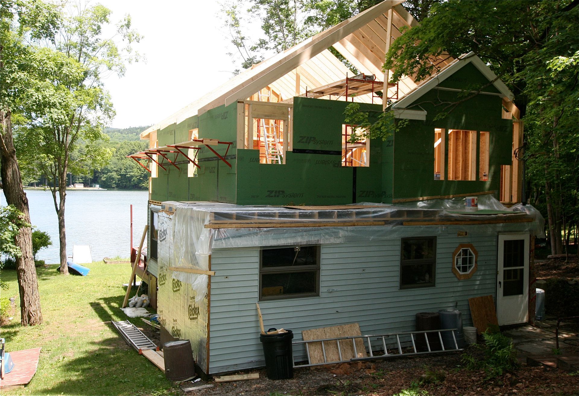 Two-story lakeside cabin under construction, with green siding and exposed roof framing amid trees.