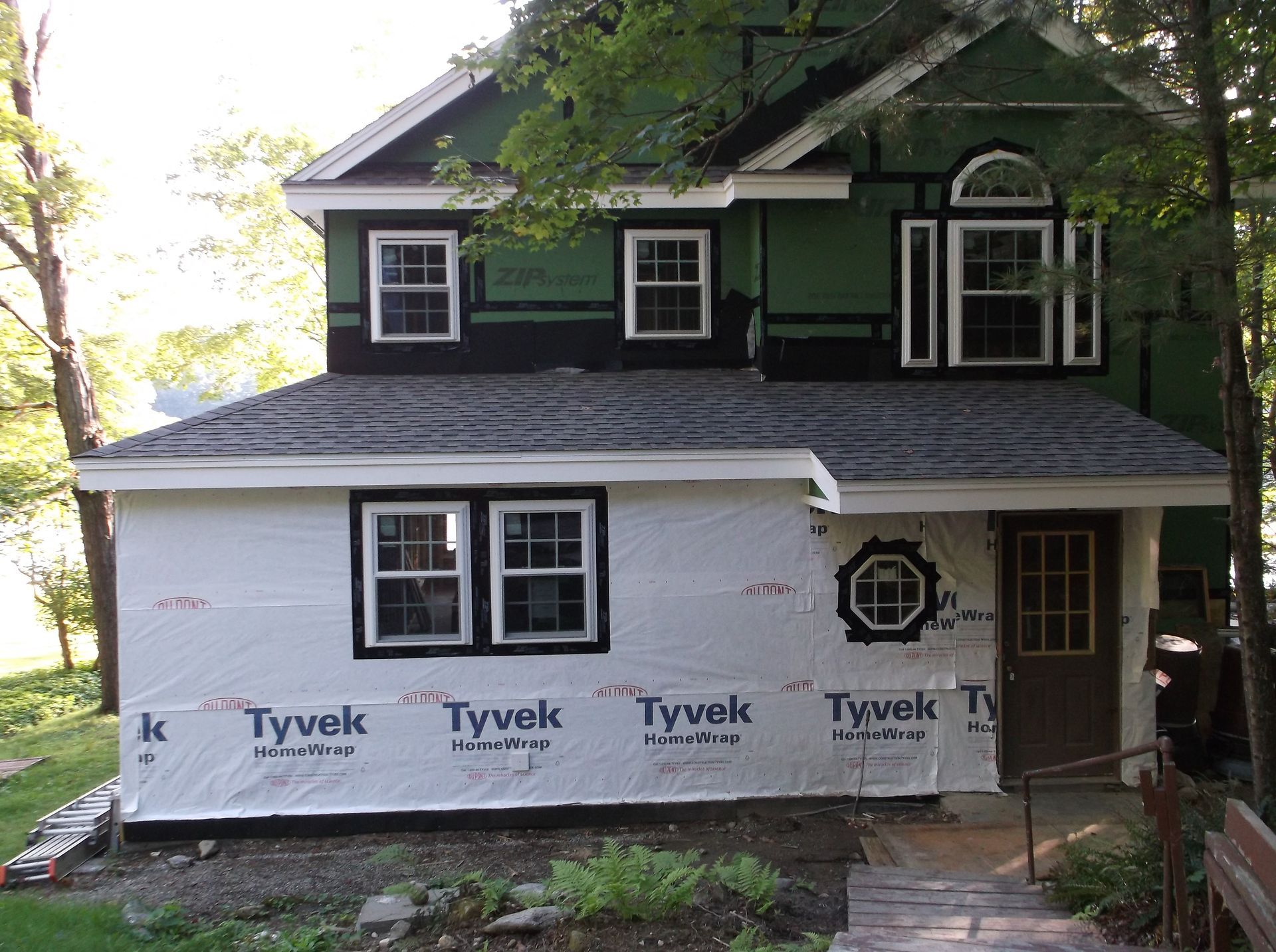 Two-story house with dark green siding and white Tyvek wrap on the lower front under renovation.