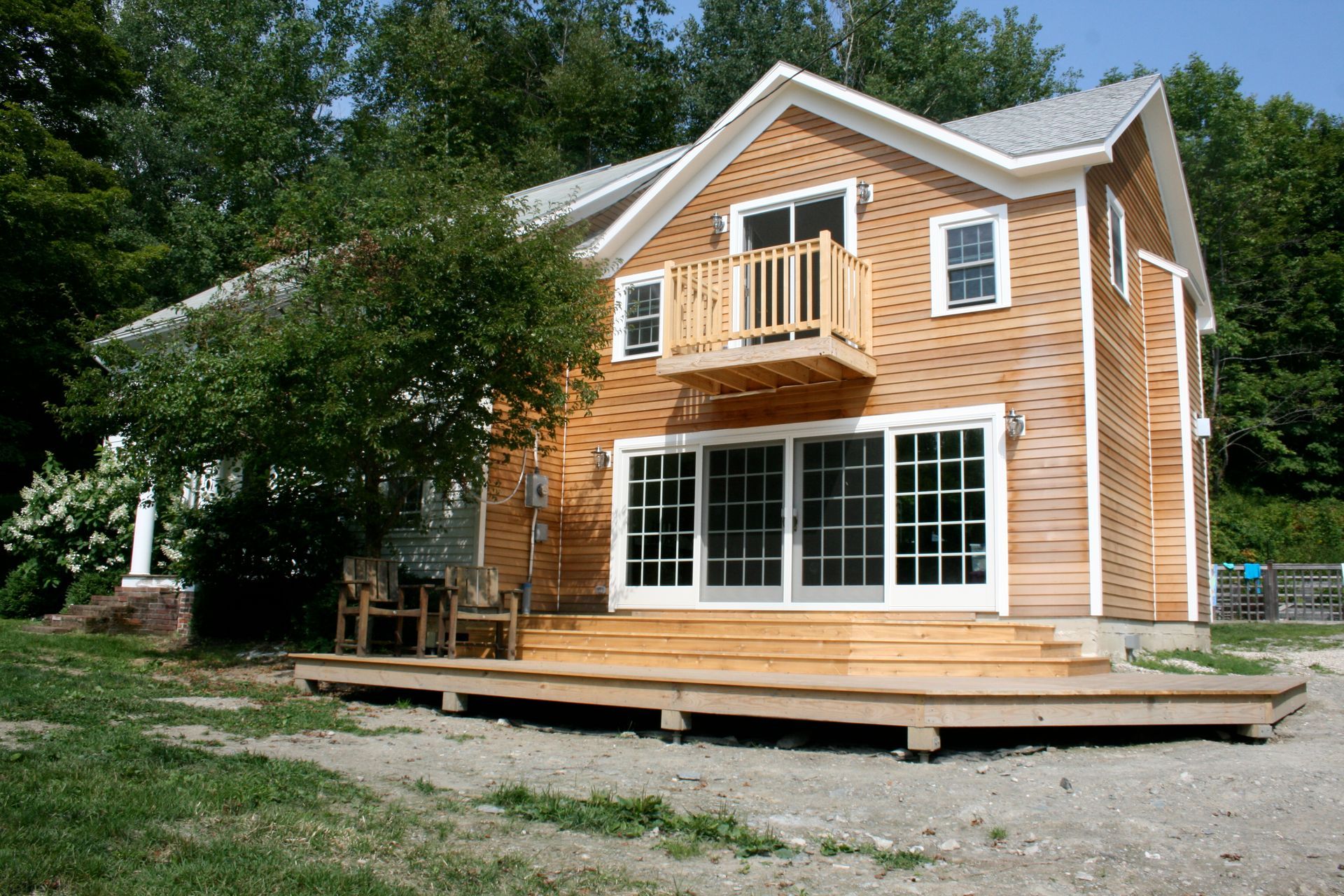 Two-story wooden house with a large deck and white-trimmed windows, surrounded by trees.