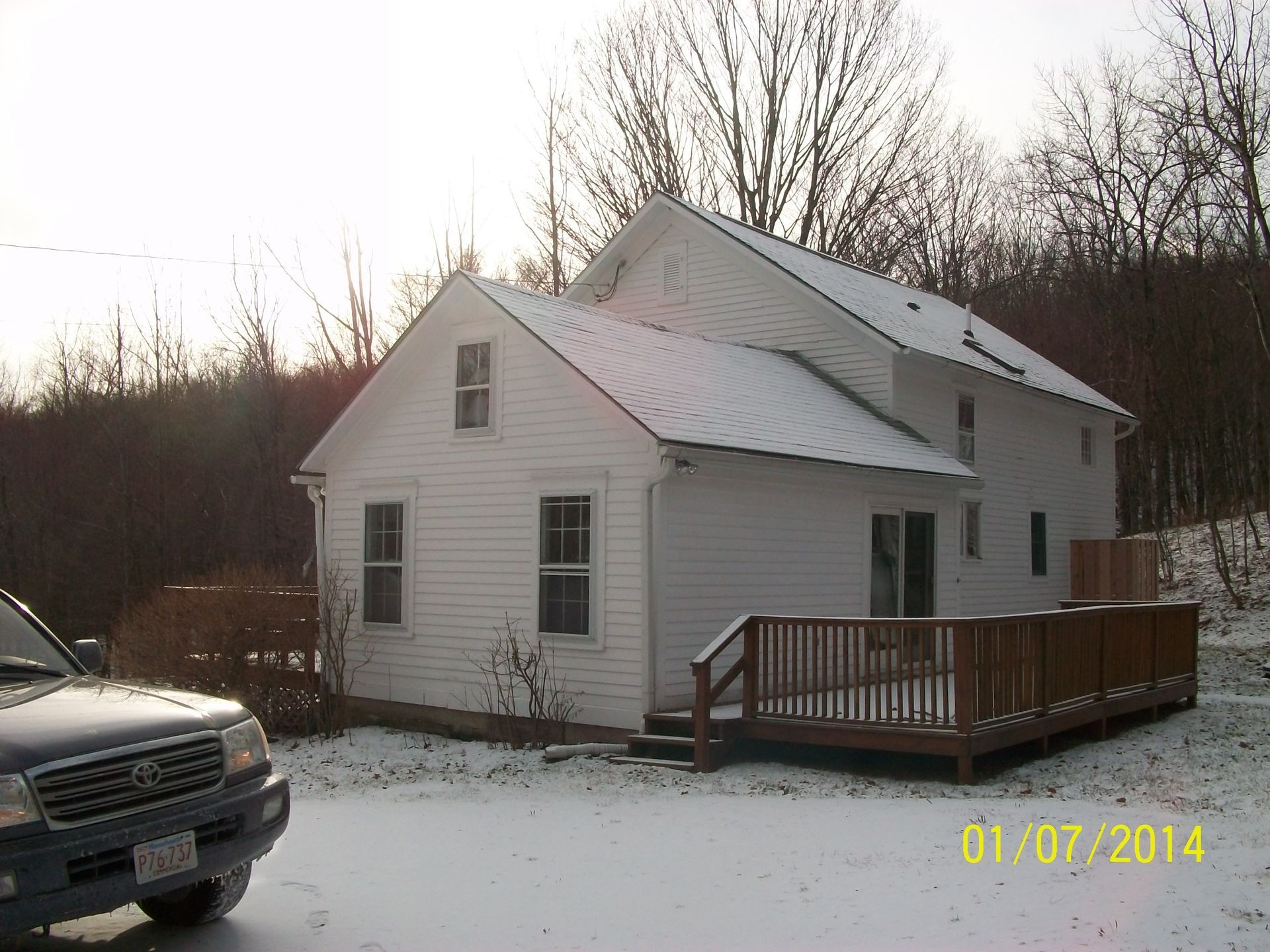 Snowy yard with a white house and wooden deck, with a car parked in the foreground.