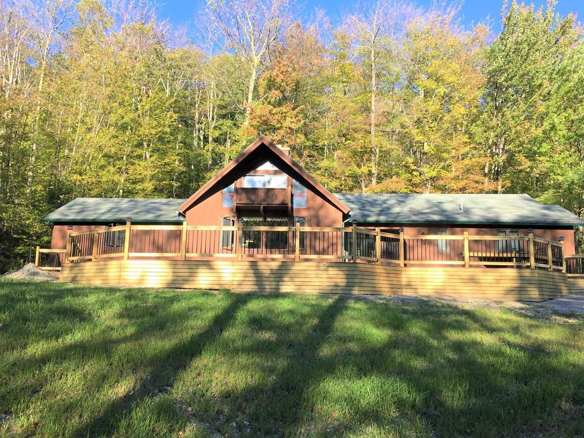 Wooden cabin with a long porch in front of autumn trees and a grassy lawn.
