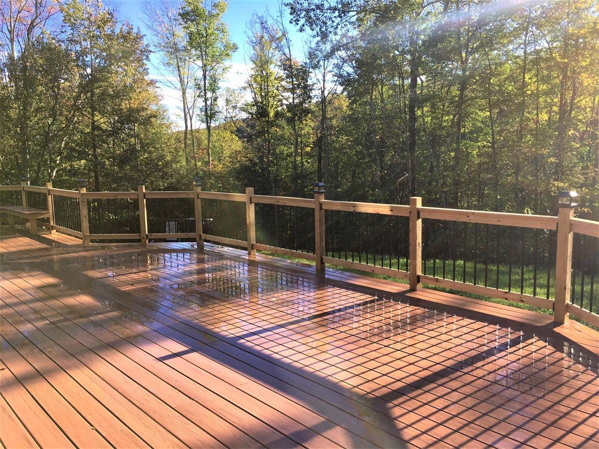 Sunlit wooden deck with a railing and trees in the background, with a wet patch reflecting the sky.