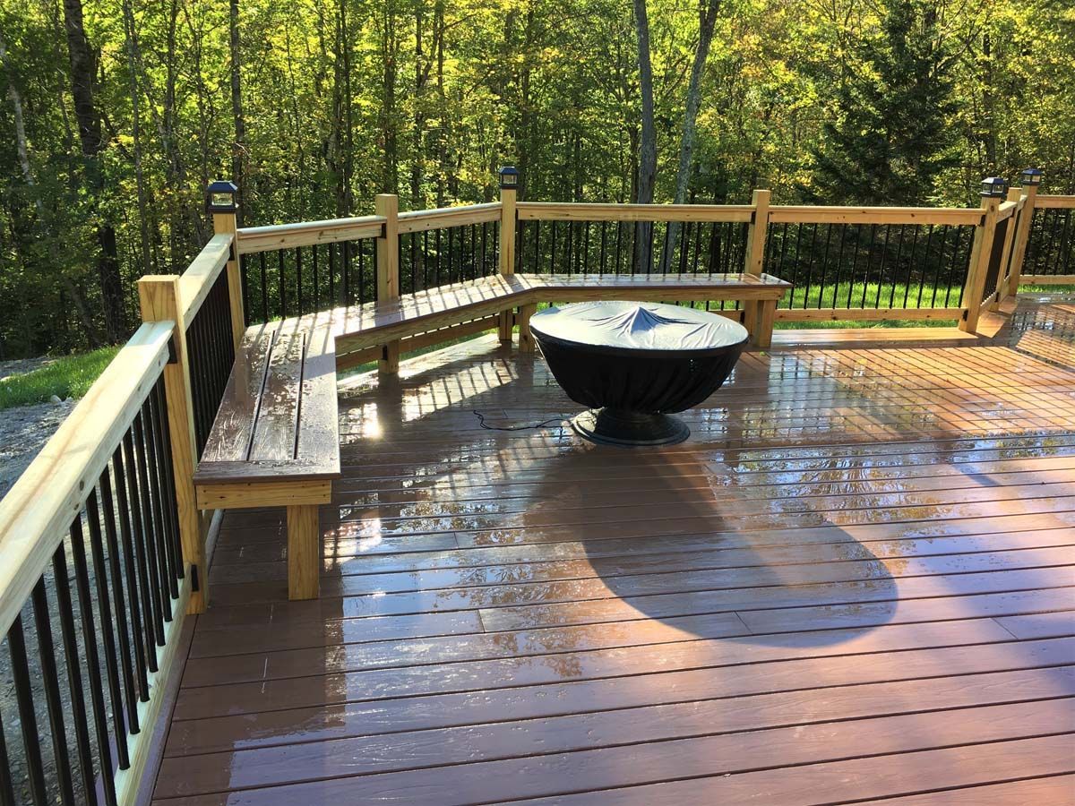 Wooden deck with bench, fire pit, and railing overlooking a green wooded area.
