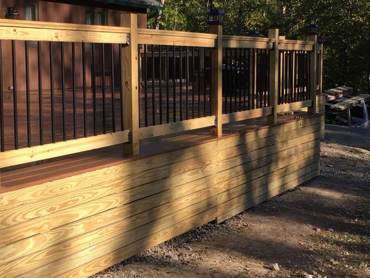 Wooden deck railing with black metal balusters in warm sunlight beside a gravel path.