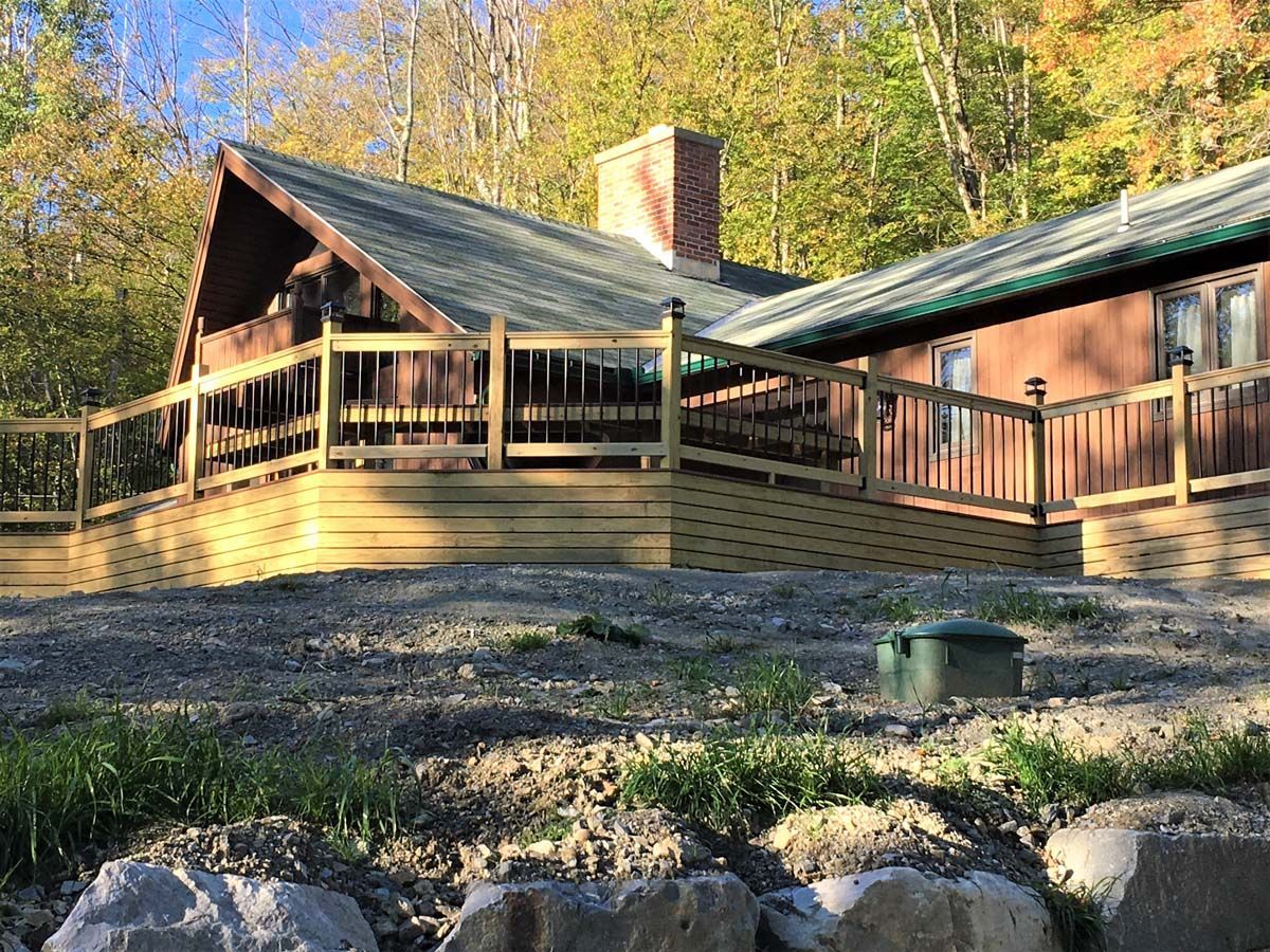 Wooden cabin with a wraparound deck in a wooded area, lit by warm sunlight.