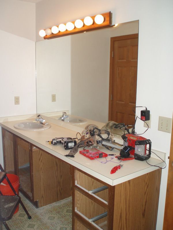 Bathroom vanity with two sinks, exposed tools and wires, and lit bulbs above a mirror.