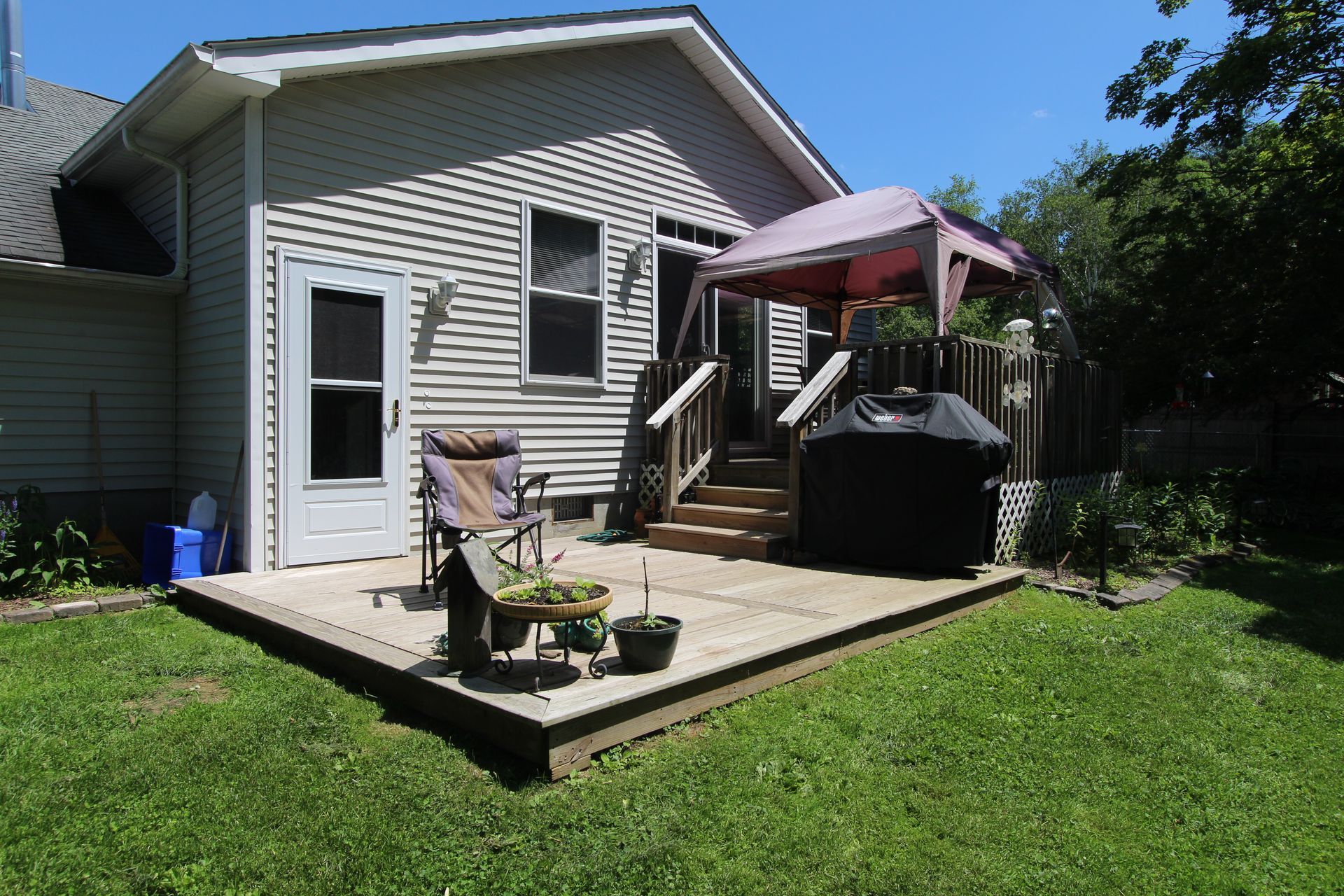 Backyard patio with grill, chairs, and potted plants beside a house on a sunny day