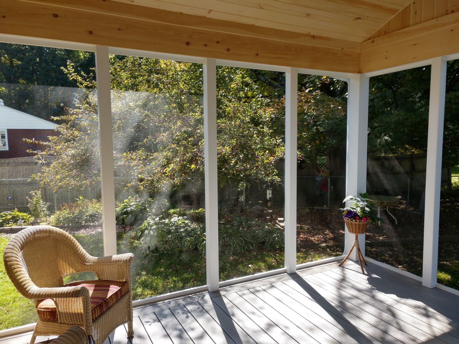Sunlit screened porch with wicker chair, potted plant, and large windows overlooking greenery