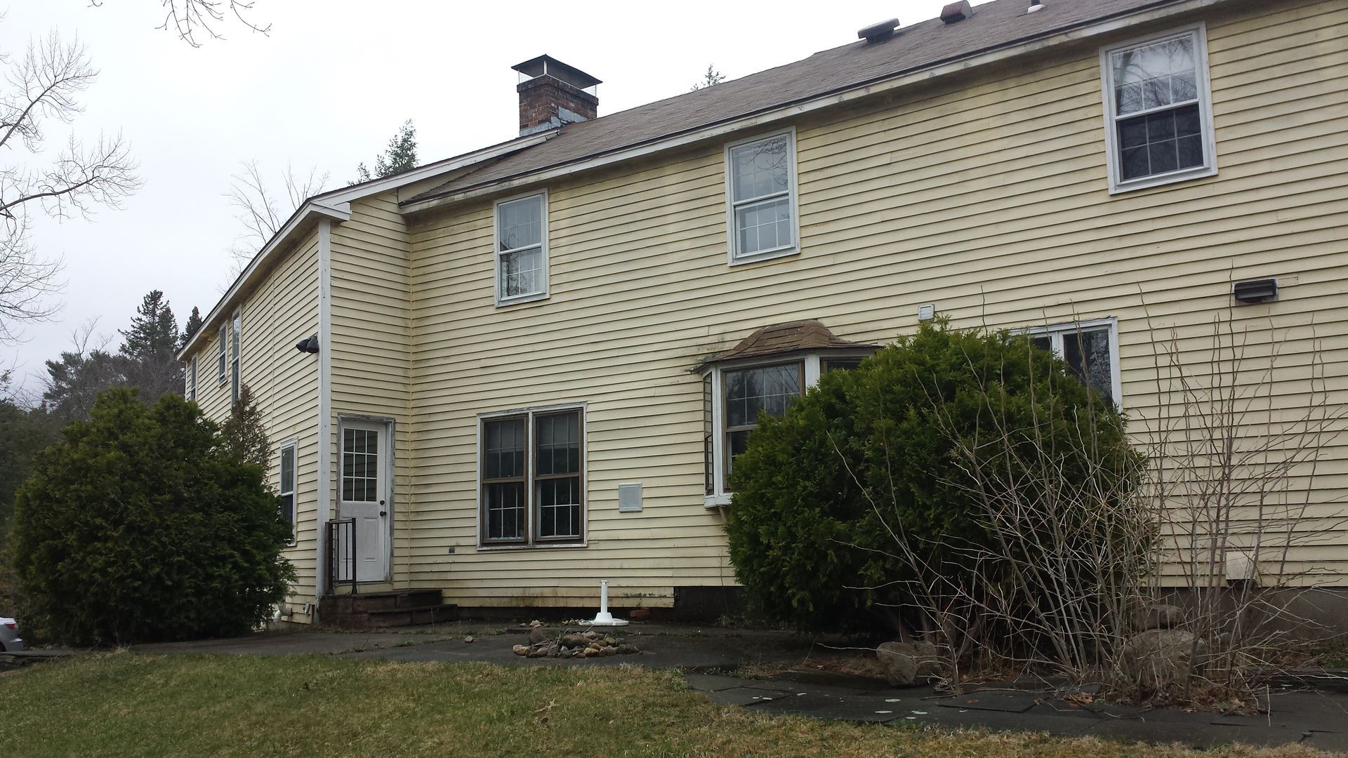 Two-story beige stone house with white-trimmed windows and bushes in a yard on an overcast day
