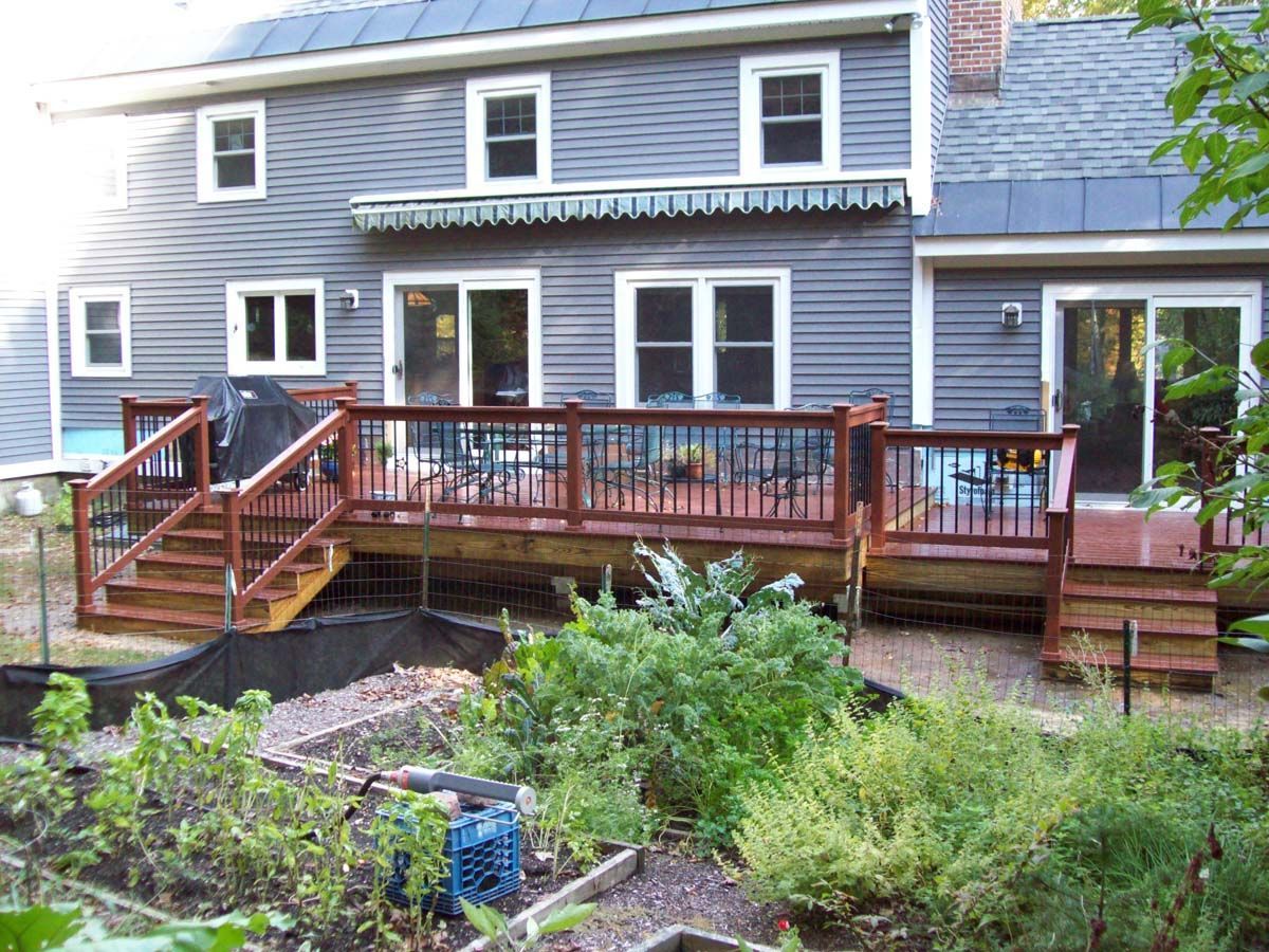 Wooden deck with railing attached to a gray house, overlooking a garden with plants and flower beds.