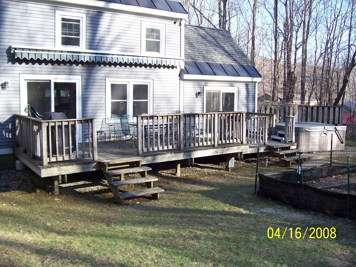Two-story house with a wooden deck and steps in a grassy yard, leafless trees in the background.