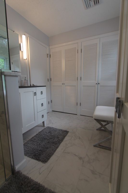 Bright bathroom with marble tile, white vanity, louvered closet doors, and a gray rug.