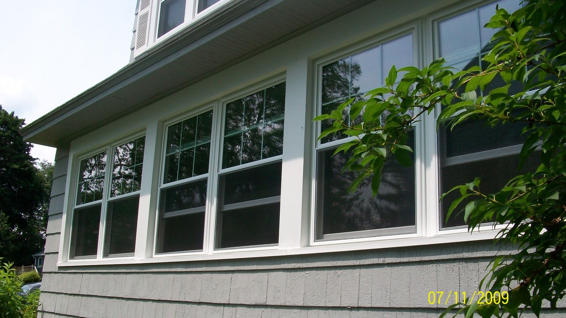White-framed house windows under a gray roof, partly shaded by green tree branches