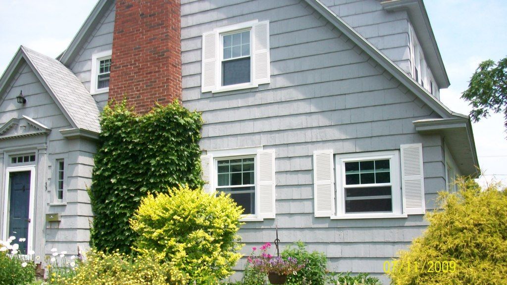 Gray house with white trim and red brick chimney, partly covered by ivy and surrounded by green shrubs.