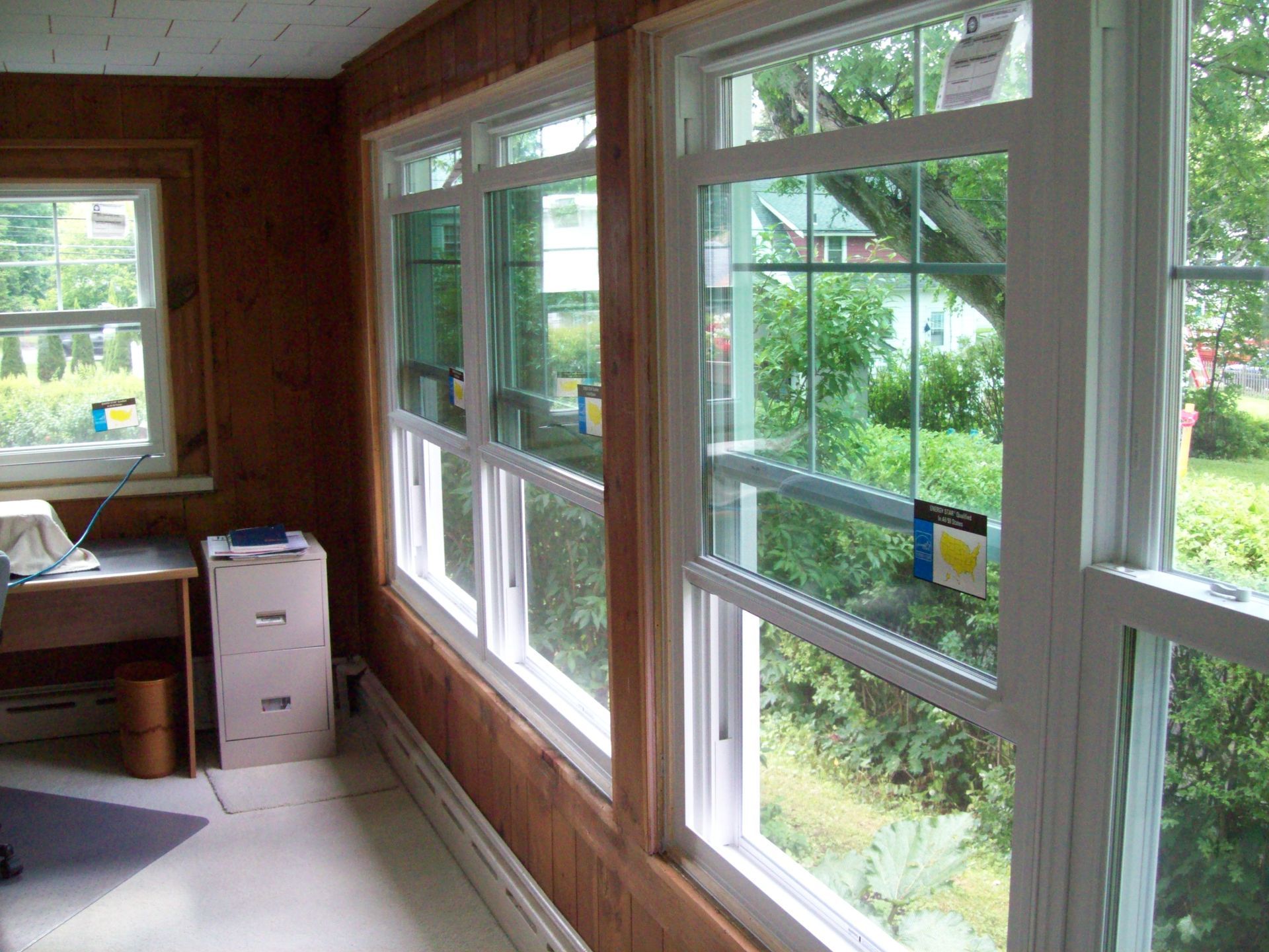 Sunlit sunroom with large windows, wood paneling, desk, and a small white cabinet by the wall.