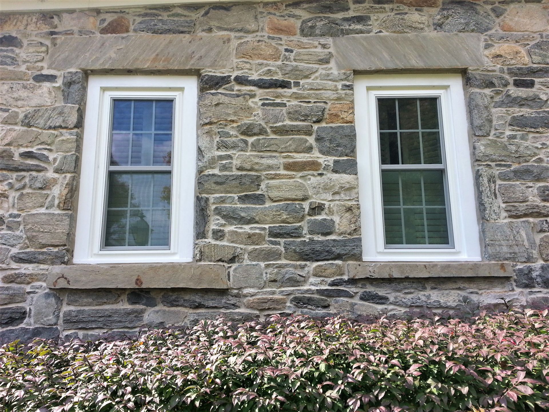 Stone house wall with two white-framed windows above pink flowers