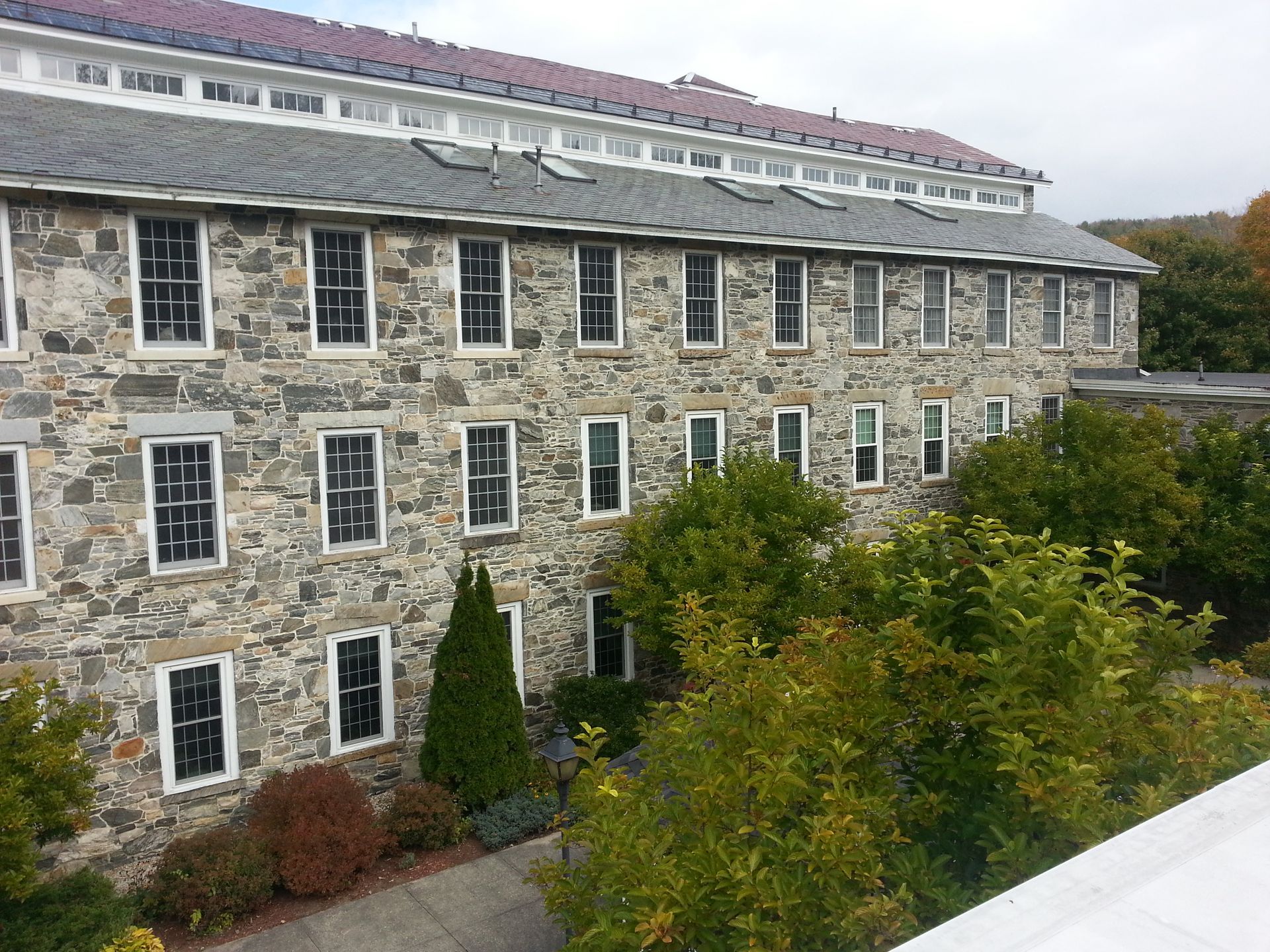 Stone building with rows of windows and a red roof, viewed from a landscaped courtyard.