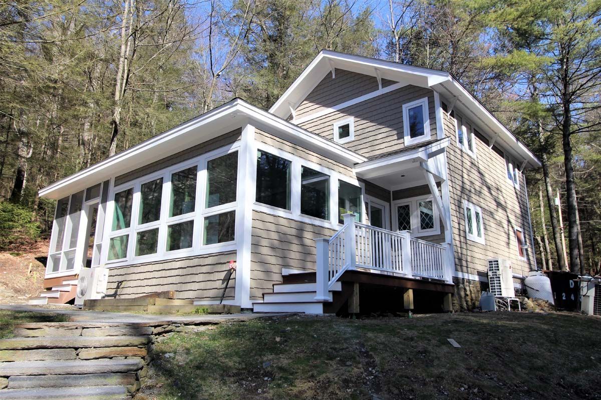 Two-story beige house with large white windows and a screened porch on a wooded hillside