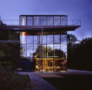 Modern glass house with a lit interior at dusk, viewed from the front.
