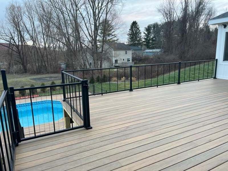 Wooden deck with black railing overlooking a backyard pool and leafless trees