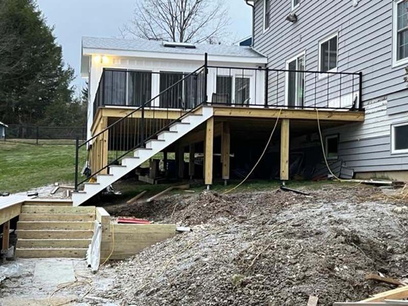 House deck under construction with stairs, black railings, and exposed supports on a sloped yard.