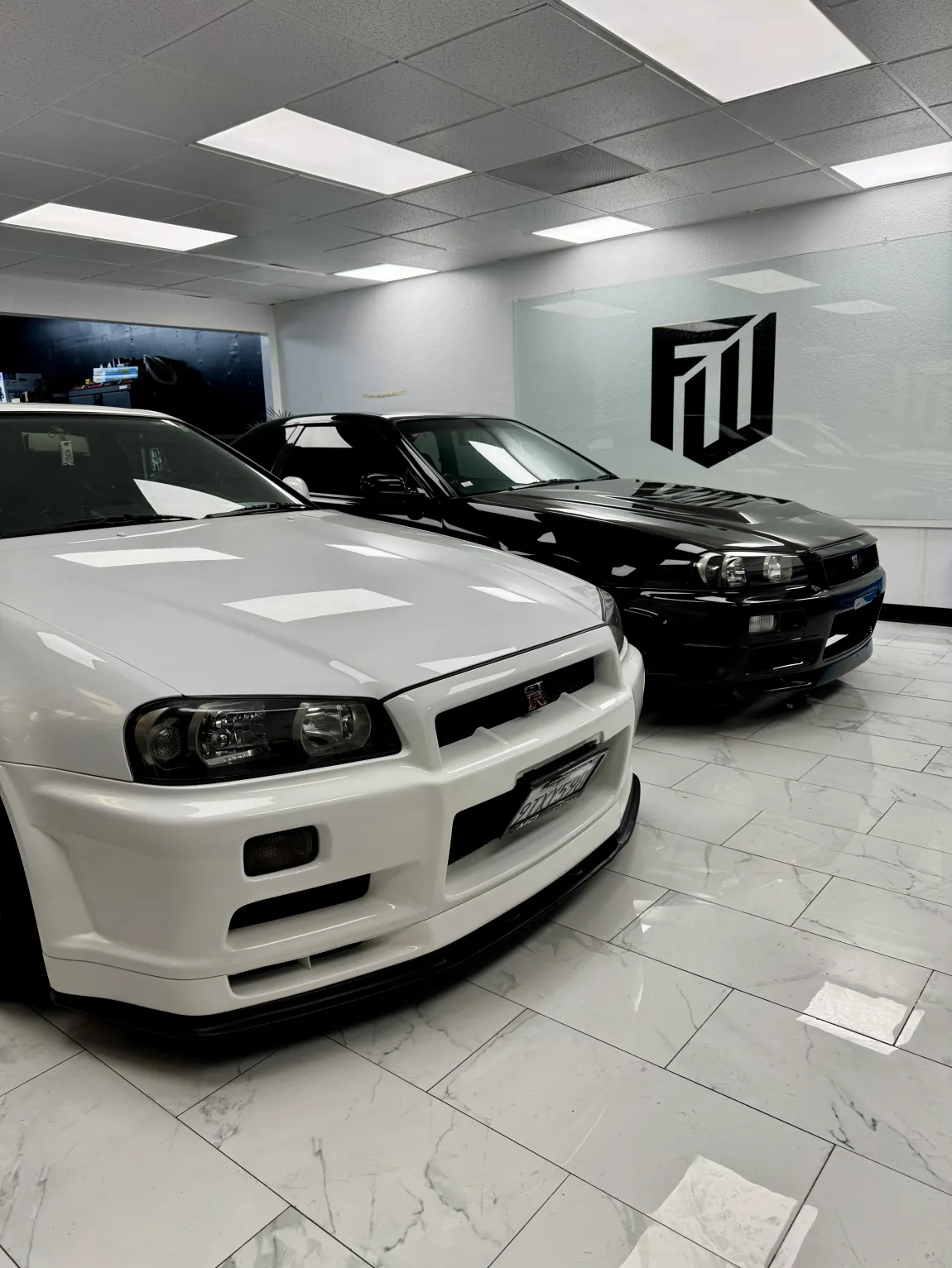 Two sports cars, white and black, displayed in a bright showroom with tiled floor.