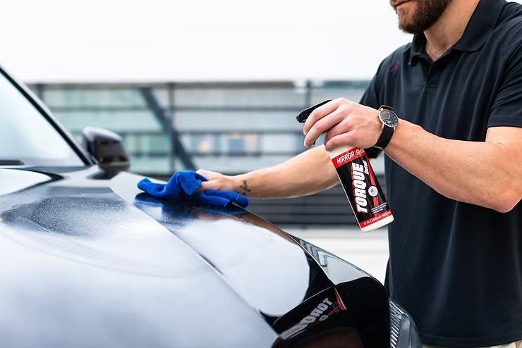 Person spraying a black car hood with cleaner and wiping it with a blue cloth