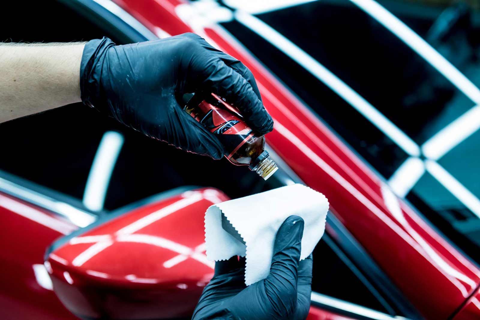 Gloved hands polishing a red car with a cloth and detailing spray under bright lights