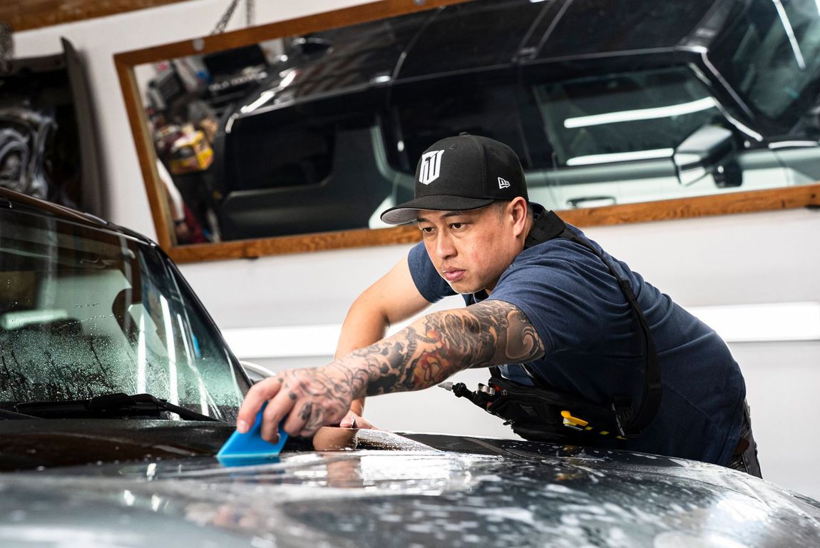 Person washing a car with a blue sponge in a garage, leaning over the hood.