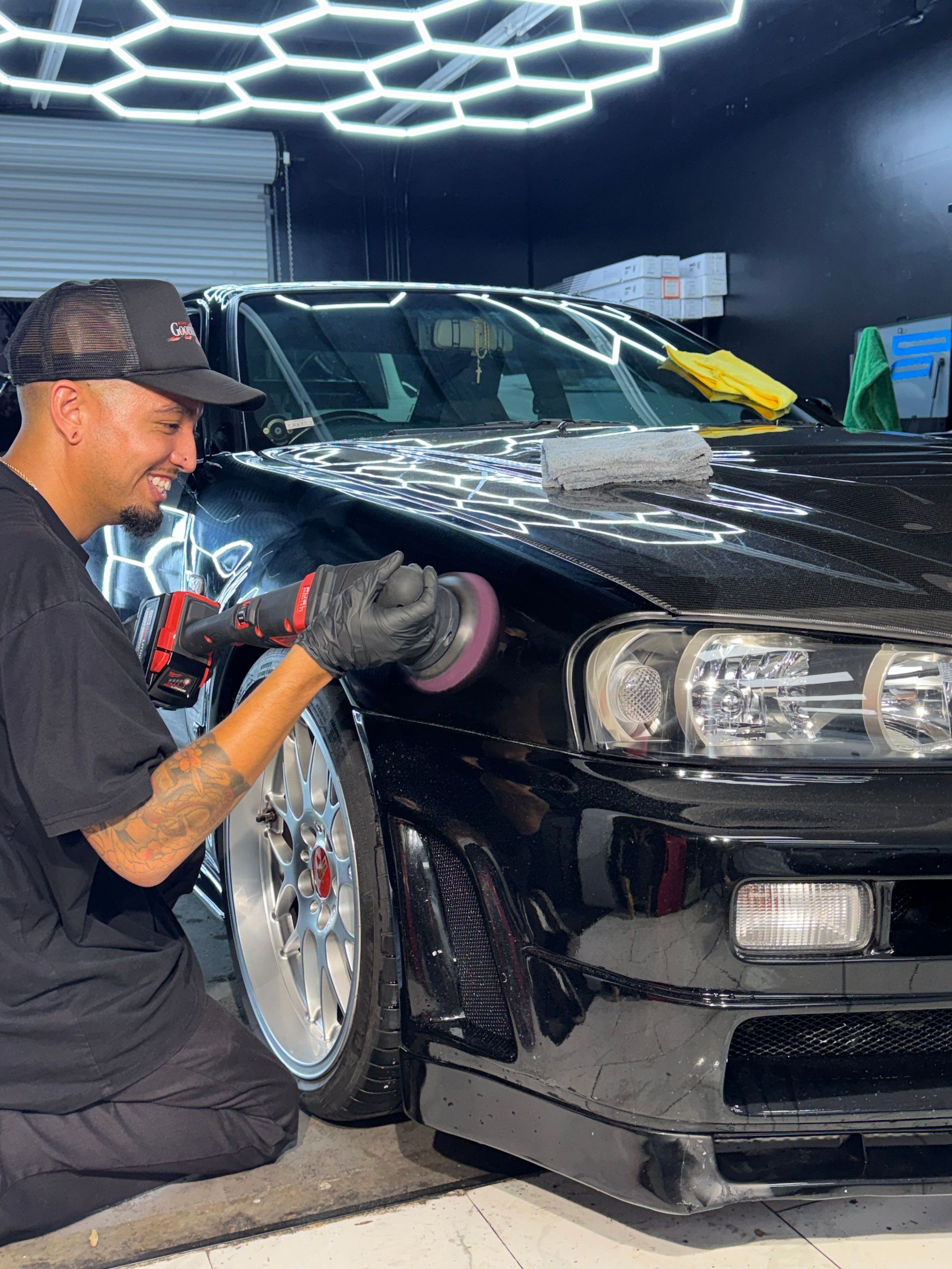 Detailing a black sports car as a man polishes the front fender in a garage, with LED ceiling lights overhead
