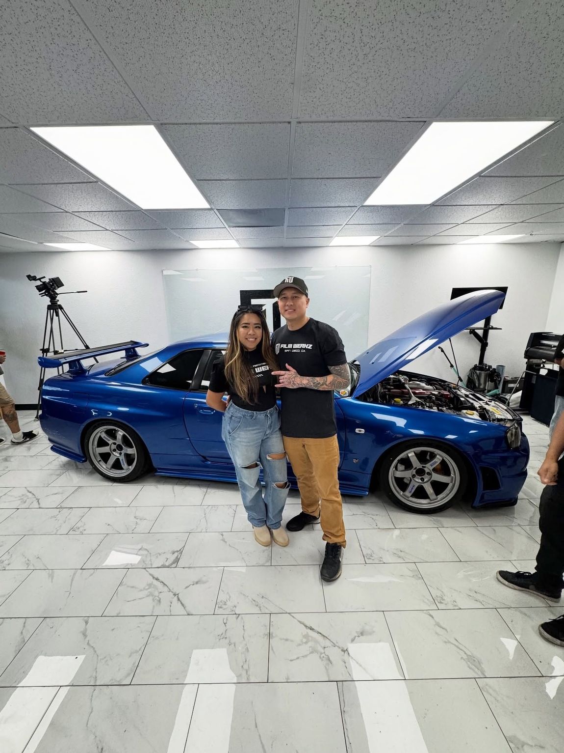 Two people posing in front of a blue car with the hood open in a bright garage