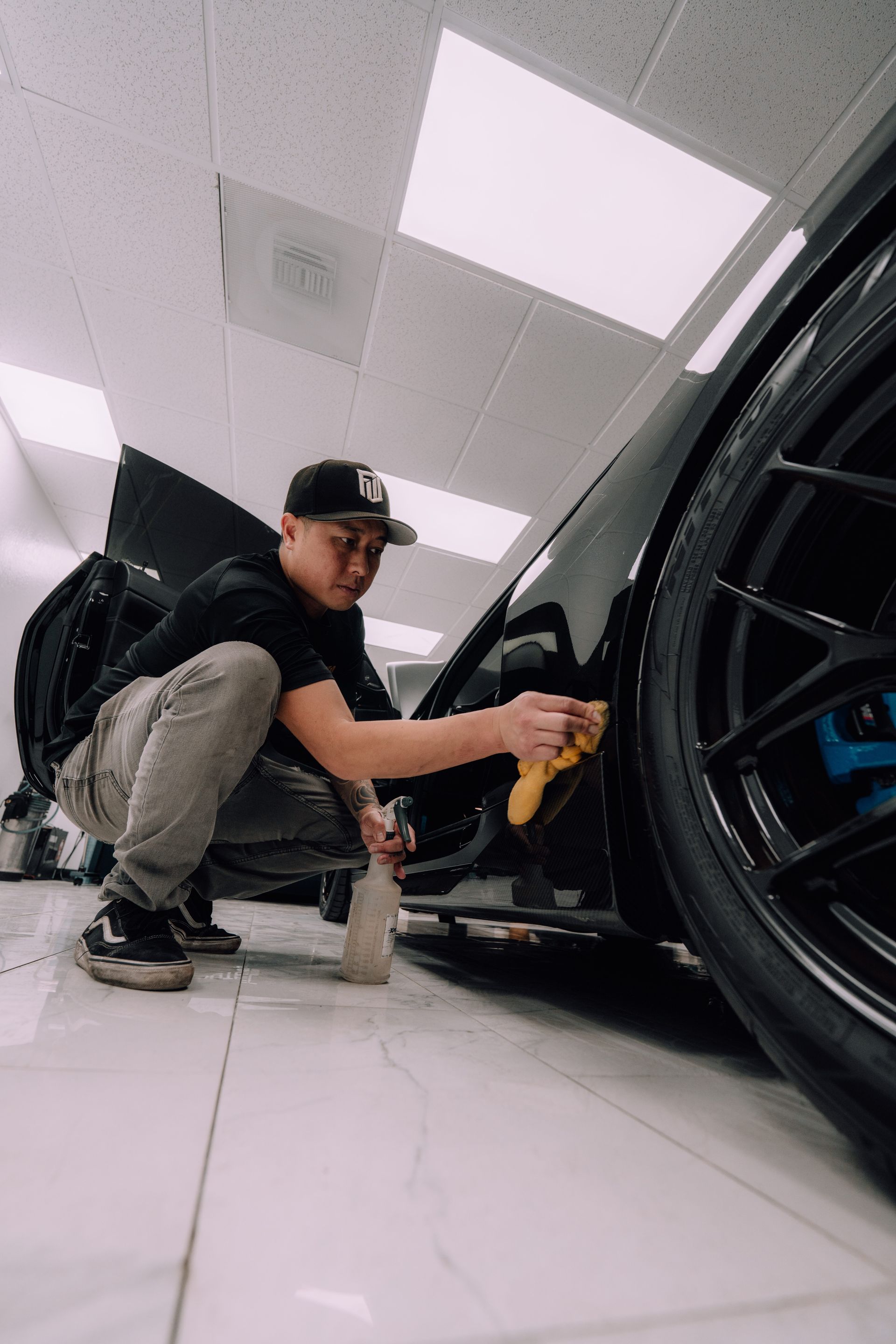 Person crouches beside a black sports car in a bright garage, cleaning the wheel with a cloth.