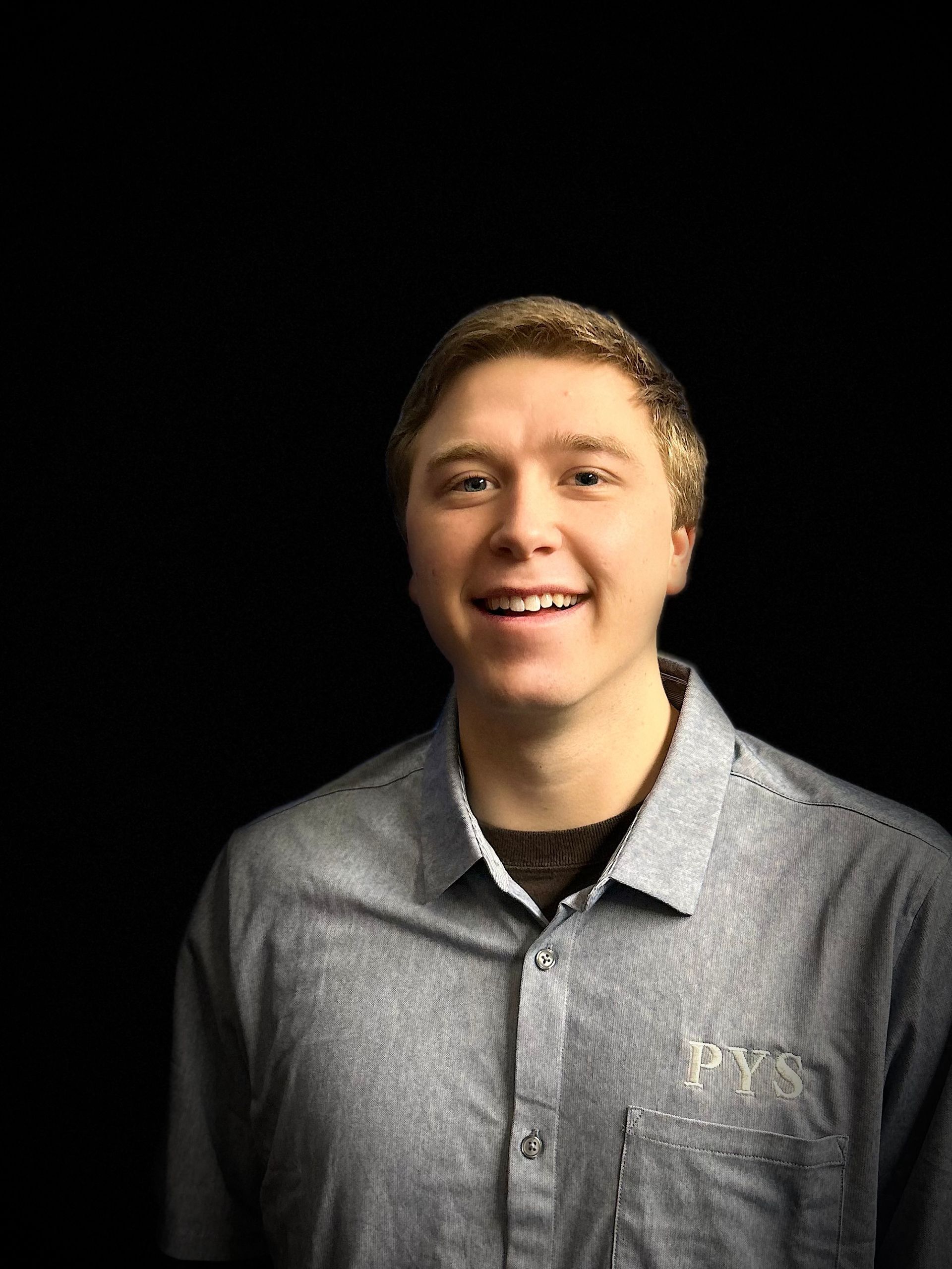 A young man in a grey shirt is smiling in front of a black background.
