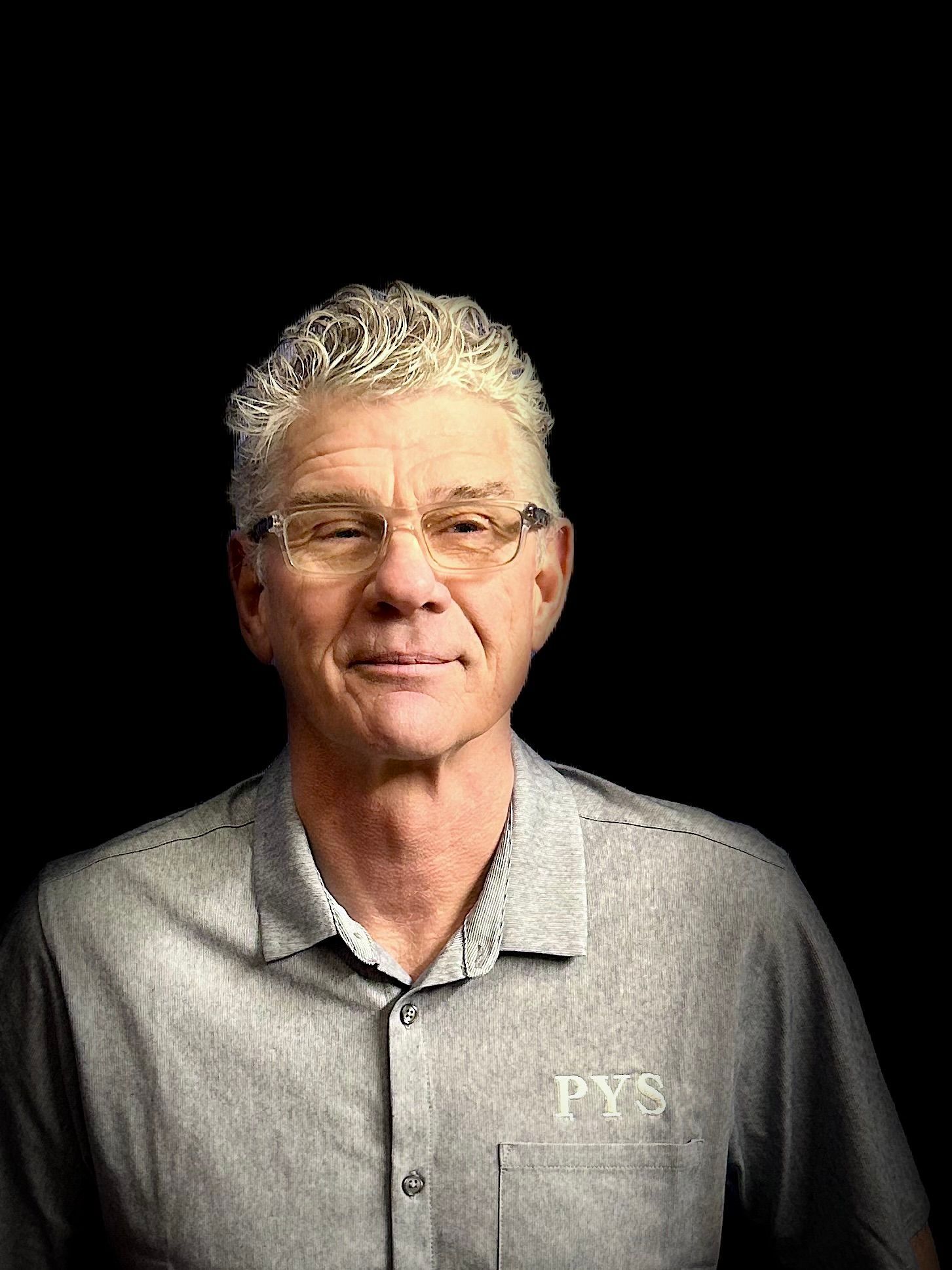 A man wearing glasses and a grey shirt is standing in front of a black background.