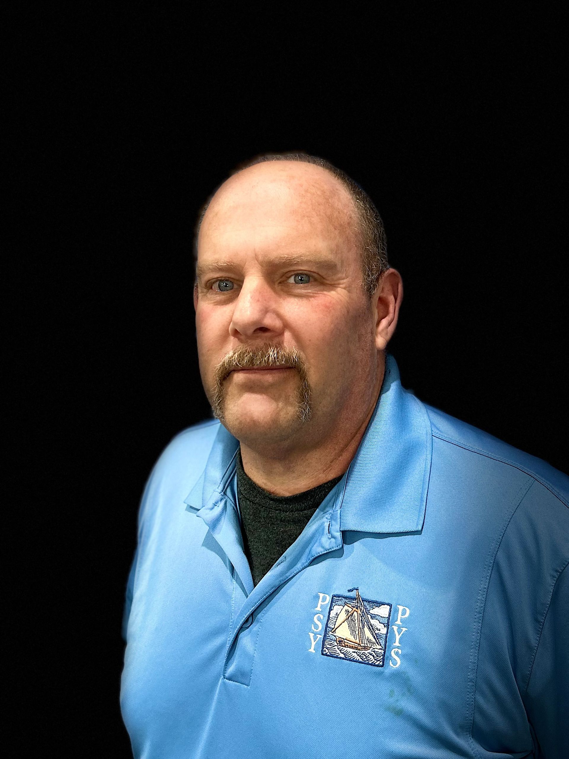 A man with a mustache and a blue shirt is standing in front of a black background.