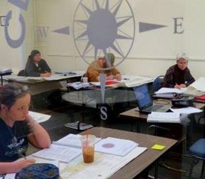 A group of people are sitting at tables in a classroom with a compass on the wall.